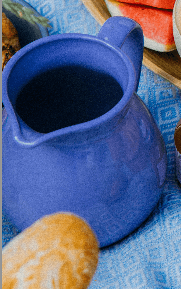 Glossy periwinkle-blue ceramic pitcher on a blue patterned tablecloth, with bread and sliced watermelon around it