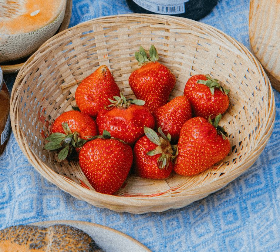 Shallow woven basket of ripe red strawberries on a blue patterned picnic cloth, next to a halved melon