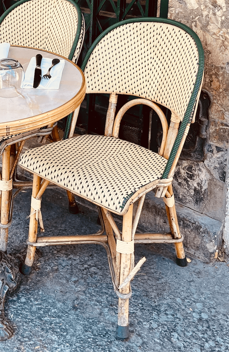 Classic Parisian bistro chair of rattan with a cream-and-black woven seat and green trim, next to a round marble table set with a knife, fork and napkin