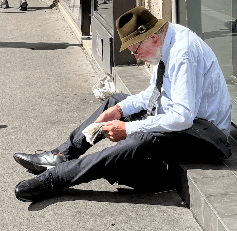 Older man with a white beard, a brown felt hat, glasses, a light blue shirt and a tie sitting on a sunlit sidewalk step reading a folded newspaper